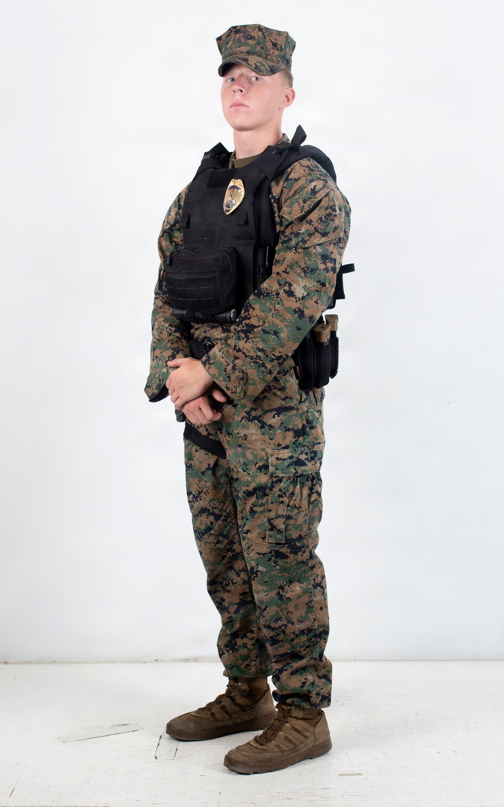A young male individual stands in a three-quarter pose against a plain white background. He is wearing woodland digital camouflage (MARPAT-pattern) uniform with a black tactical vest/plate carrier that displays a gold badge on the chest. The image appears to be a professional reference or documentation photograph.