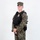 MCAS Marines pose for content 2026 A young male individual stands in a three-quarter pose against a plain white background. He is wearing woodland digital camouflage (MARPAT-pattern) uniform with a black tactical vest/plate carrier that displays a gold badge on the chest. The image appears to be a professional reference or documentation photograph.
