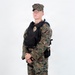 A young male individual stands in a three-quarter pose against a plain white background. He is wearing woodland digital camouflage (MARPAT-pattern) uniform with a black tactical vest/plate carrier that displays a gold badge on the chest. The image appears to be a professional reference or documentation photograph.