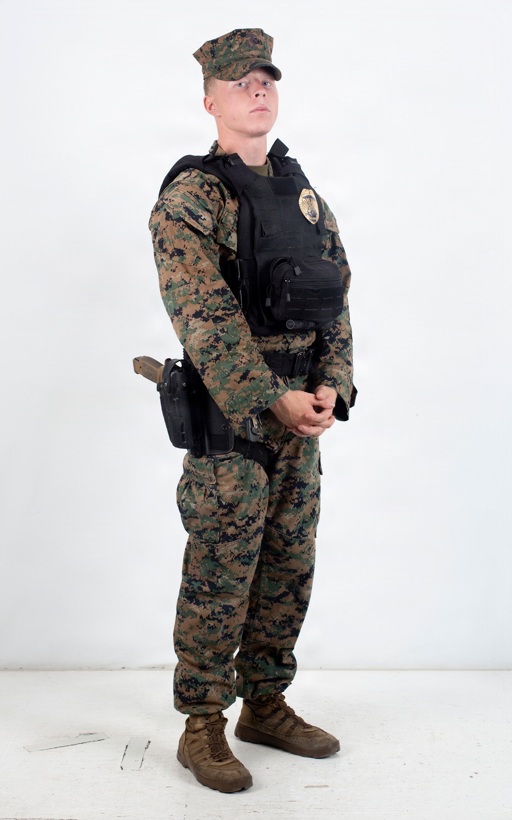 A young male in USMC MARPAT woodland digital camouflage uniform stands in a neutral pose against a white studio background. He is wearing a black tactical plate carrier vest with a visible gold badge, a black duty belt with a thigh holster containing a pistol, and coyote brown combat boots. The image appears to be a reference or documentation photograph of military/security personnel equipment.