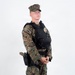 A young male in USMC MARPAT woodland digital camouflage uniform stands in a neutral pose against a white studio background. He is wearing a black tactical plate carrier vest with a visible gold badge, a black duty belt with a thigh holster containing a pistol, and coyote brown combat boots. The image appears to be a reference or documentation photograph of military/security personnel equipment.