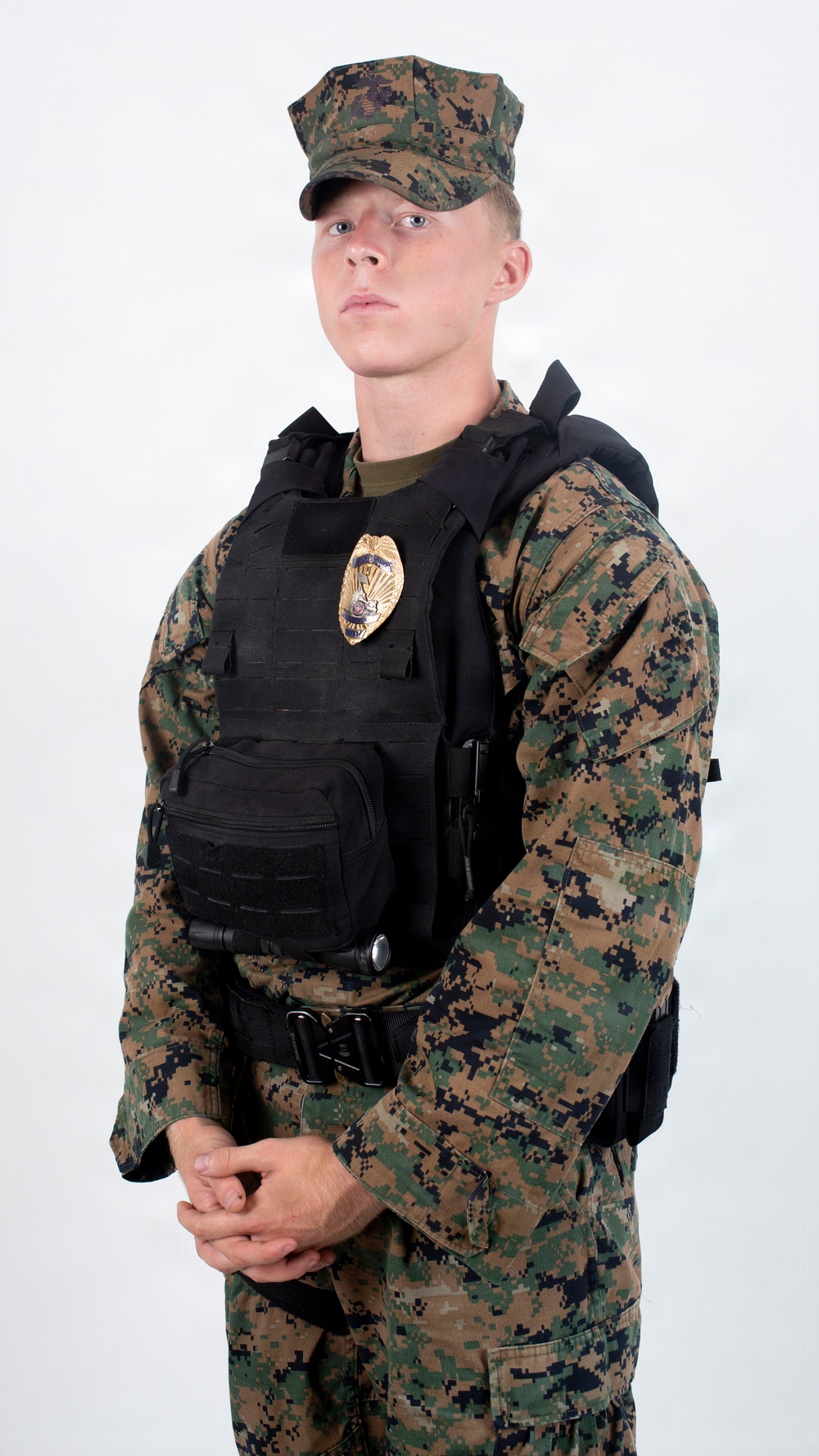 A young male in MARPAT (Marine Pattern) woodland digital camouflage uniform is posed against a plain white background. He is wearing a black tactical plate carrier vest with a gold law enforcement badge affixed to the front, along with MOLLE-compatible pouches. The image appears to be a professional or studio-style portrait, likely for documentation or display purposes.
