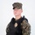 MCAS Marines pose for content 2026 A young male in MARPAT (Marine Pattern) woodland digital camouflage uniform is posed against a plain white background. He is wearing a black tactical plate carrier vest with a gold law enforcement badge affixed to the front, along with MOLLE-compatible pouches. The image appears to be a professional or studio-style portrait, likely for documentation or display purposes.