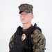 A young male in MARPAT (Marine Pattern) woodland digital camouflage uniform is posed against a plain white background. He is wearing a black tactical plate carrier vest with a gold law enforcement badge affixed to the front, along with MOLLE-compatible pouches. The image appears to be a professional or studio-style portrait, likely for documentation or display purposes.