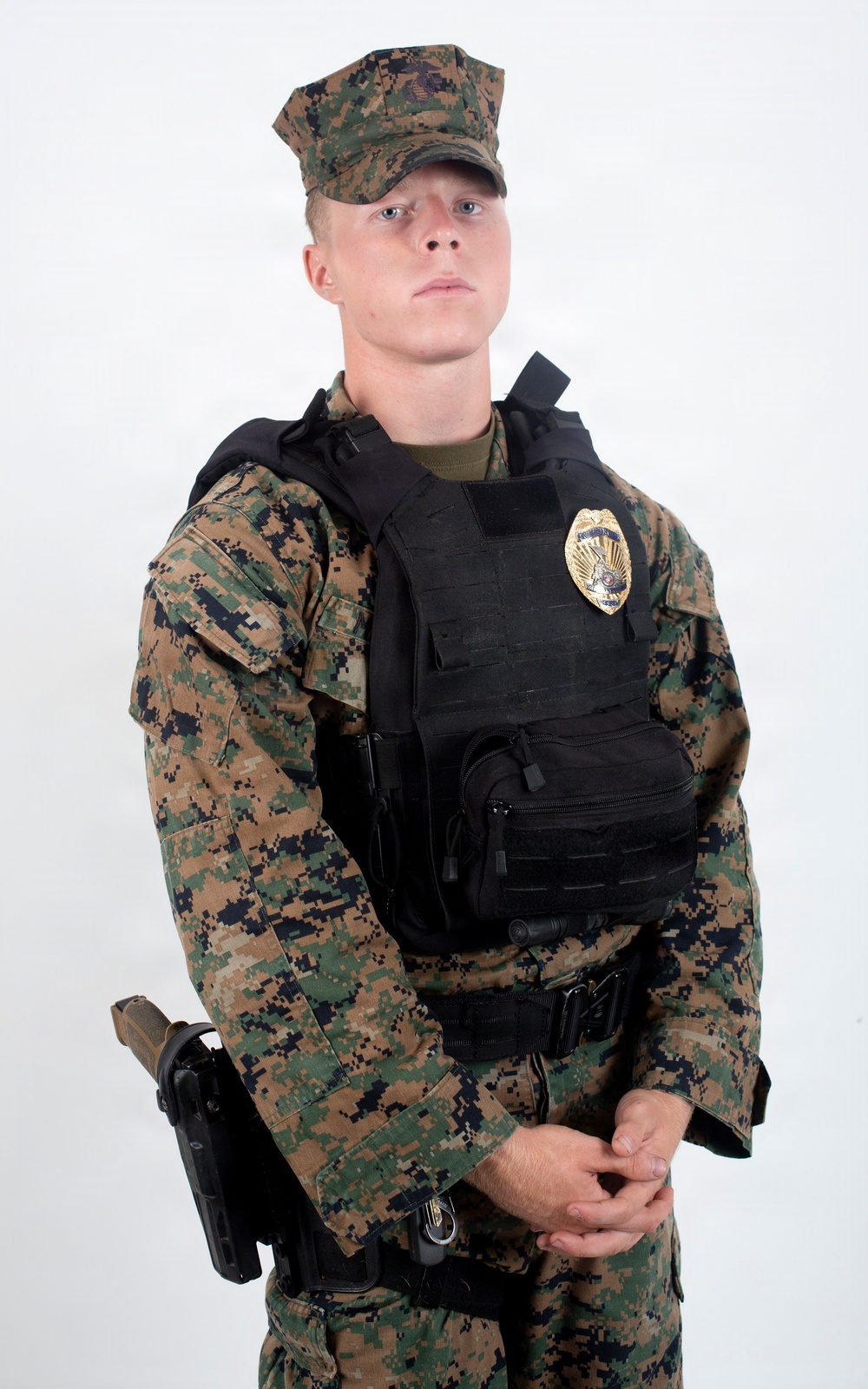 A young male in USMC MARPAT woodland digital camouflage uniform wearing a black tactical plate carrier vest with a gold law enforcement badge affixed to the chest. He is equipped with a duty belt and a holstered sidearm on his left hip. The image appears to be a professional portrait or official documentation photo taken against a plain white background.