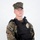 MCAS Marines pose for content 2026 A young male in USMC MARPAT woodland digital camouflage uniform wearing a black tactical plate carrier vest with a gold law enforcement badge affixed to the chest. He is equipped with a duty belt and a holstered sidearm on his left hip. The image appears to be a professional portrait or official documentation photo taken against a plain white background.
