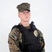 A young male in USMC MARPAT woodland digital camouflage uniform wearing a black tactical plate carrier vest with a gold law enforcement badge affixed to the chest. He is equipped with a duty belt and a holstered sidearm on his left hip. The image appears to be a professional portrait or official documentation photo taken against a plain white background.