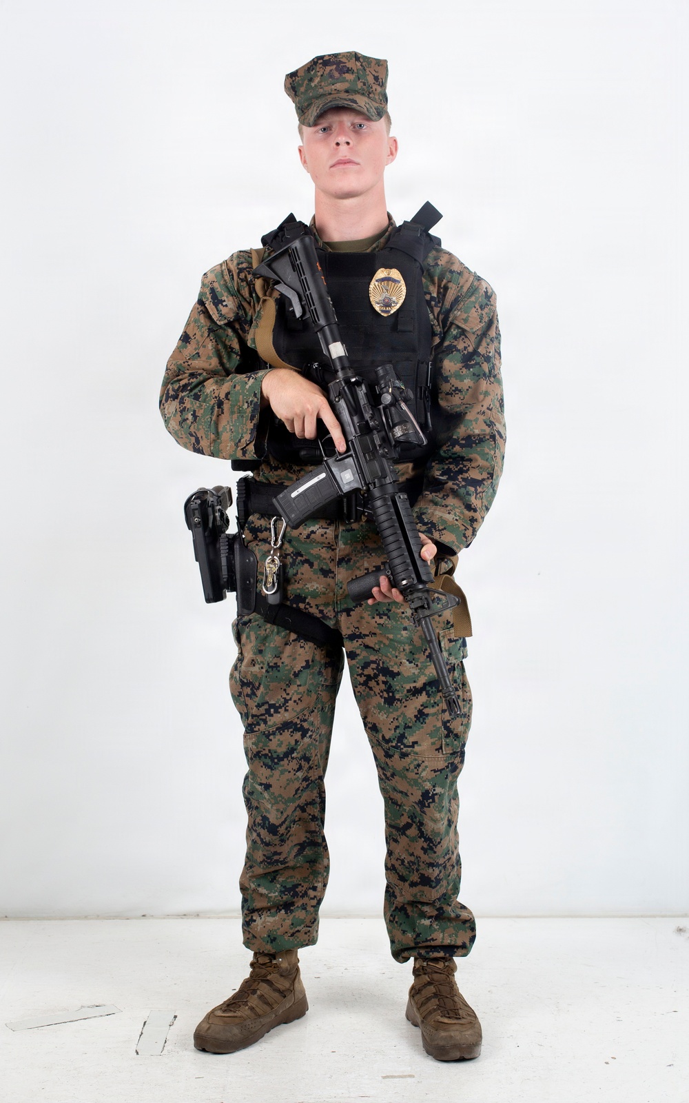 A young male in USMC MARPAT woodland digital camouflage uniform stands in a studio setting against a white background. He is wearing a black tactical vest with a visible gold badge, and is holding an AR-style rifle. His kit includes a holstered sidearm and additional duty belt equipment.