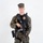 MCAS Marines pose for content 2026 A young male in USMC MARPAT woodland digital camouflage uniform stands in a studio setting against a white background. He is wearing a black tactical vest with a visible gold badge, and is holding an AR-style rifle. His kit includes a holstered sidearm and additional duty belt equipment.