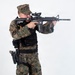 A male figure wearing USMC MARPAT woodland digital camouflage uniform is depicted in a shooting stance, aiming a rifle toward the right of the frame. The subject is equipped with full tactical gear including body armor, a holstered sidearm, and various pouches. The image appears to be a professional studio or controlled-environment photograph against a plain white background.