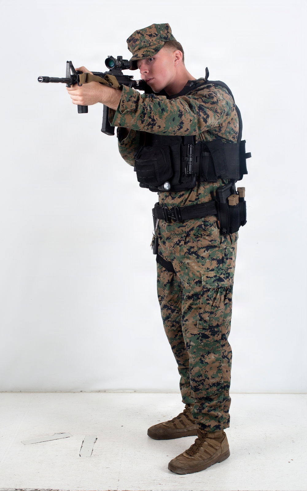 A male figure in U.S. Marine Corps MARPAT woodland digital camouflage uniform is photographed against a plain white studio background. He is wearing full tactical gear including a black plate carrier/vest with MOLLE pouches and is aiming a rifle with an optical scope. The image appears to be a professional equipment or training reference photograph.