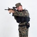 A male figure in U.S. Marine Corps MARPAT woodland digital camouflage uniform is photographed against a plain white studio background. He is wearing full tactical gear including a black plate carrier/vest with MOLLE pouches and is aiming a rifle with an optical scope. The image appears to be a professional equipment or training reference photograph.