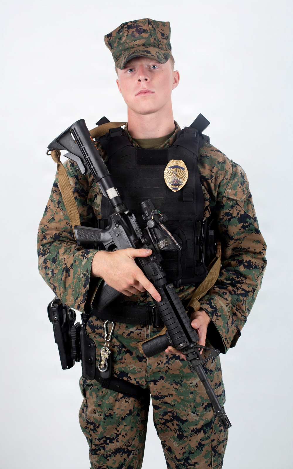 A young male in MARPAT (Marine pattern) digital camouflage uniform and cap is posed against a plain white background. He is wearing a black tactical plate carrier vest with a gold law enforcement badge affixed to it, and is holding an AR-style rifle with an optic and vertical foregrip. The combination of military camouflage and a police/security badge suggests a military police or base security role.