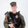 MCAS Marines pose for content 2026 A young male in MARPAT (Marine pattern) digital camouflage uniform and cap is posed against a plain white background. He is wearing a black tactical plate carrier vest with a gold law enforcement badge affixed to it, and is holding an AR-style rifle with an optic and vertical foregrip. The combination of military camouflage and a police/security badge suggests a military police or base security role.