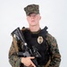 A young male in MARPAT (Marine pattern) digital camouflage uniform and cap is posed against a plain white background. He is wearing a black tactical plate carrier vest with a gold law enforcement badge affixed to it, and is holding an AR-style rifle with an optic and vertical foregrip. The combination of military camouflage and a police/security badge suggests a military police or base security role.