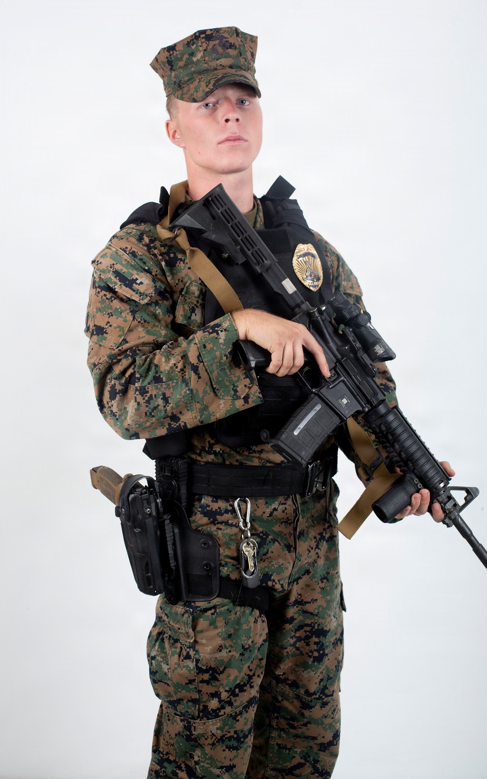 A young male in USMC MARPAT (woodland digital camouflage) uniform and cover (cap) is posed against a plain white background. He is equipped with a law enforcement or security badge, body armor, a tactical belt with holster, and is holding an M4-style carbine rifle with a tan sling. The studio-style lighting and neutral background suggest this is an official or promotional photograph.