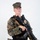 MCAS Marines pose for content 2026 A young male in USMC MARPAT (woodland digital camouflage) uniform and cover (cap) is posed against a plain white background. He is equipped with a law enforcement or security badge, body armor, a tactical belt with holster, and is holding an M4-style carbine rifle with a tan sling. The studio-style lighting and neutral background suggest this is an official or promotional photograph.