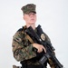 A young male in USMC MARPAT (woodland digital camouflage) uniform and cover (cap) is posed against a plain white background. He is equipped with a law enforcement or security badge, body armor, a tactical belt with holster, and is holding an M4-style carbine rifle with a tan sling. The studio-style lighting and neutral background suggest this is an official or promotional photograph.