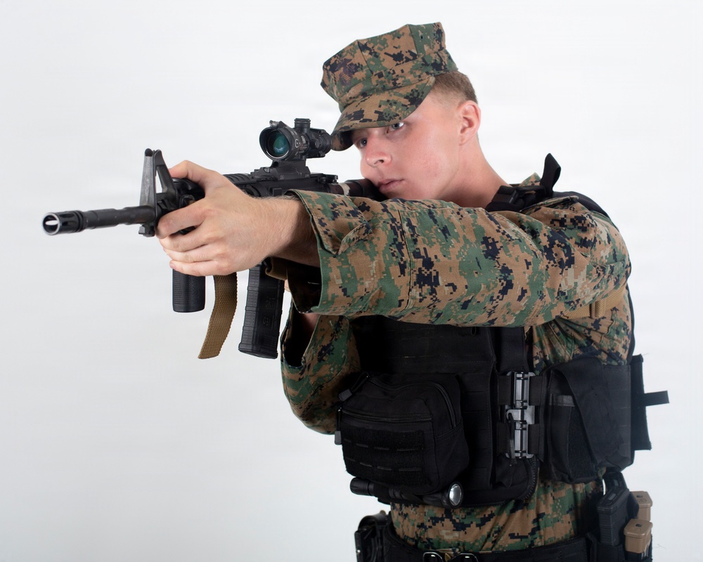 A male individual wearing USMC MARPAT woodland digital camouflage uniform and cover (cap) is posed in an aiming stance with a rifle. He is equipped with tactical body armor/plate carrier with attached pouches and accessories. The image appears to be a studio or controlled environment photograph against a plain white background.