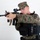MCAS Marines pose for content 2026 A male individual wearing USMC MARPAT woodland digital camouflage uniform and cover (cap) is posed in an aiming stance with a rifle. He is equipped with tactical body armor/plate carrier with attached pouches and accessories. The image appears to be a studio or controlled environment photograph against a plain white background.