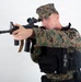 A male individual wearing USMC MARPAT woodland digital camouflage uniform and cover (cap) is posed in an aiming stance with a rifle. He is equipped with tactical body armor/plate carrier with attached pouches and accessories. The image appears to be a studio or controlled environment photograph against a plain white background.