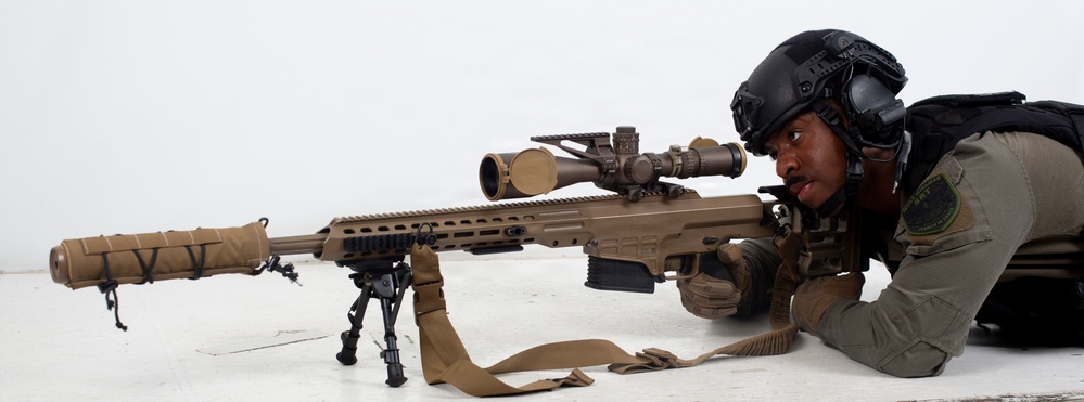 A military or law enforcement operator is in a prone shooting position aiming a large-caliber sniper rifle with a suppressor and bipod. The individual is wearing tactical gear including a ballistic helmet, body armor, and gloves. The setting appears to be an outdoor or rooftop environment with a light-colored surface.
