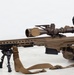 A military or law enforcement operator is in a prone shooting position aiming a large-caliber sniper rifle with a suppressor and bipod. The individual is wearing tactical gear including a ballistic helmet, body armor, and gloves. The setting appears to be an outdoor or rooftop environment with a light-colored surface.