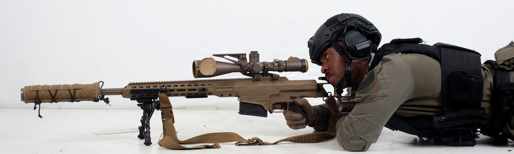 A military or law enforcement sniper is shown in a prone shooting position, aiming a large-caliber precision rifle equipped with a high-powered scope. The individual is wearing full tactical gear including a ballistic helmet, hearing protection, body armor, and tactical gloves. The image appears to be a professional or training photograph taken against a neutral background.