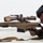 MCAS Marines pose for content 2026 A military or law enforcement sniper is shown in a prone shooting position, aiming a large-caliber precision rifle equipped with a high-powered scope. The individual is wearing full tactical gear including a ballistic helmet, hearing protection, body armor, and tactical gloves. The image appears to be a professional or training photograph taken against a neutral background.