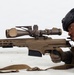 A military or law enforcement sniper is shown in a prone shooting position, aiming a large-caliber precision rifle equipped with a high-powered scope. The individual is wearing full tactical gear including a ballistic helmet, hearing protection, body armor, and tactical gloves. The image appears to be a professional or training photograph taken against a neutral background.