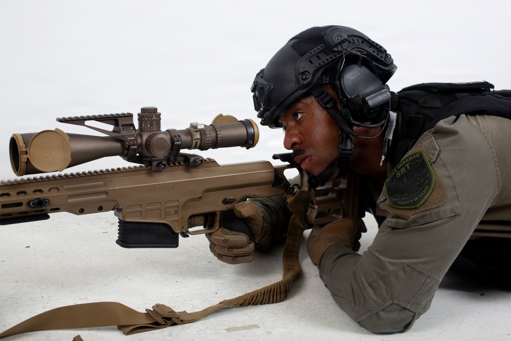 A law enforcement officer in tactical gear is in a prone shooting position behind a large precision rifle equipped with a high-powered scope. The officer is wearing a tactical helmet with hearing protection, body armor, and a uniform patch identifying 'Camp Miramar SRT' (Special Response Team). The image appears to be taken during a training exercise or demonstration.