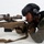 MCAS Marines pose for content 2026 A law enforcement officer in tactical gear is in a prone shooting position behind a large precision rifle equipped with a high-powered scope. The officer is wearing a tactical helmet with hearing protection, body armor, and a uniform patch identifying 'Camp Miramar SRT' (Special Response Team). The image appears to be taken during a training exercise or demonstration.