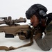 A law enforcement officer in tactical gear is in a prone shooting position behind a large precision rifle equipped with a high-powered scope. The officer is wearing a tactical helmet with hearing protection, body armor, and a uniform patch identifying 'Camp Miramar SRT' (Special Response Team). The image appears to be taken during a training exercise or demonstration.