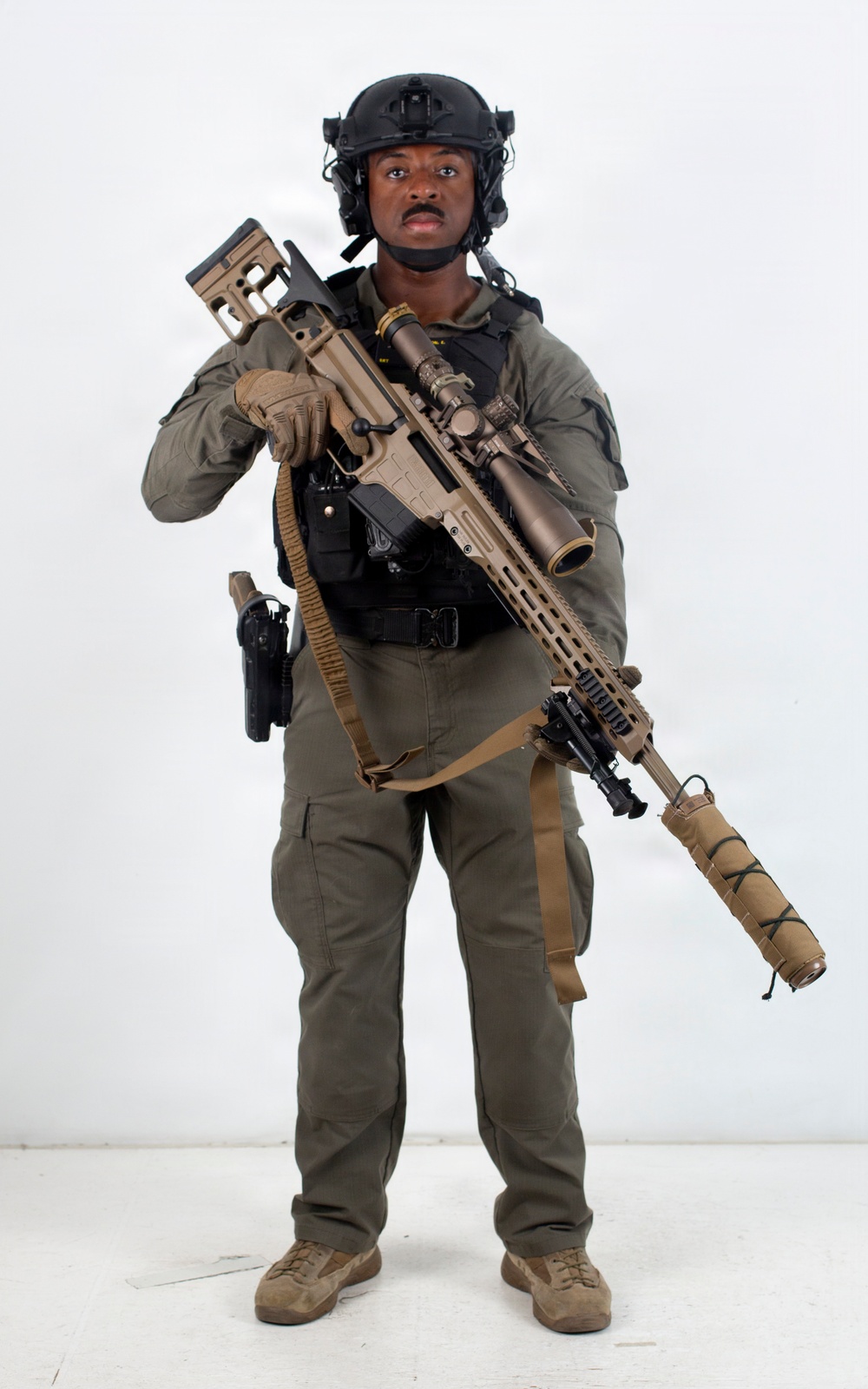 A person in full tactical/military-style gear is posed against a plain white background in what appears to be a studio or promotional photograph. They are equipped with a large precision rifle with a suppressor and scope, body armor, helmet, and sidearm. The image appears to be a professional equipment showcase or gear demonstration photo.