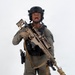 A person in full tactical/military-style gear is posed against a plain white background in what appears to be a studio or promotional photograph. They are equipped with a large precision rifle with a suppressor and scope, body armor, helmet, and sidearm. The image appears to be a professional equipment showcase or gear demonstration photo.
