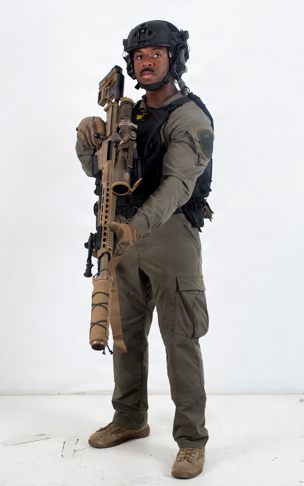 A person in full tactical law enforcement or military-style gear is posed against a plain white background in what appears to be a professional portrait or equipment showcase photo. They are equipped with a large precision rifle with a suppressor and scope, wearing a tactical vest, ballistic helmet with communications equipment, and olive drab uniform. A green patch on the right sleeve reads 'SRT' (Special Response Team).
