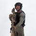 A person in full tactical law enforcement or military-style gear is posed against a plain white background in what appears to be a professional portrait or equipment showcase photo. They are equipped with a large precision rifle with a suppressor and scope, wearing a tactical vest, ballistic helmet with communications equipment, and olive drab uniform. A green patch on the right sleeve reads 'SRT' (Special Response Team).