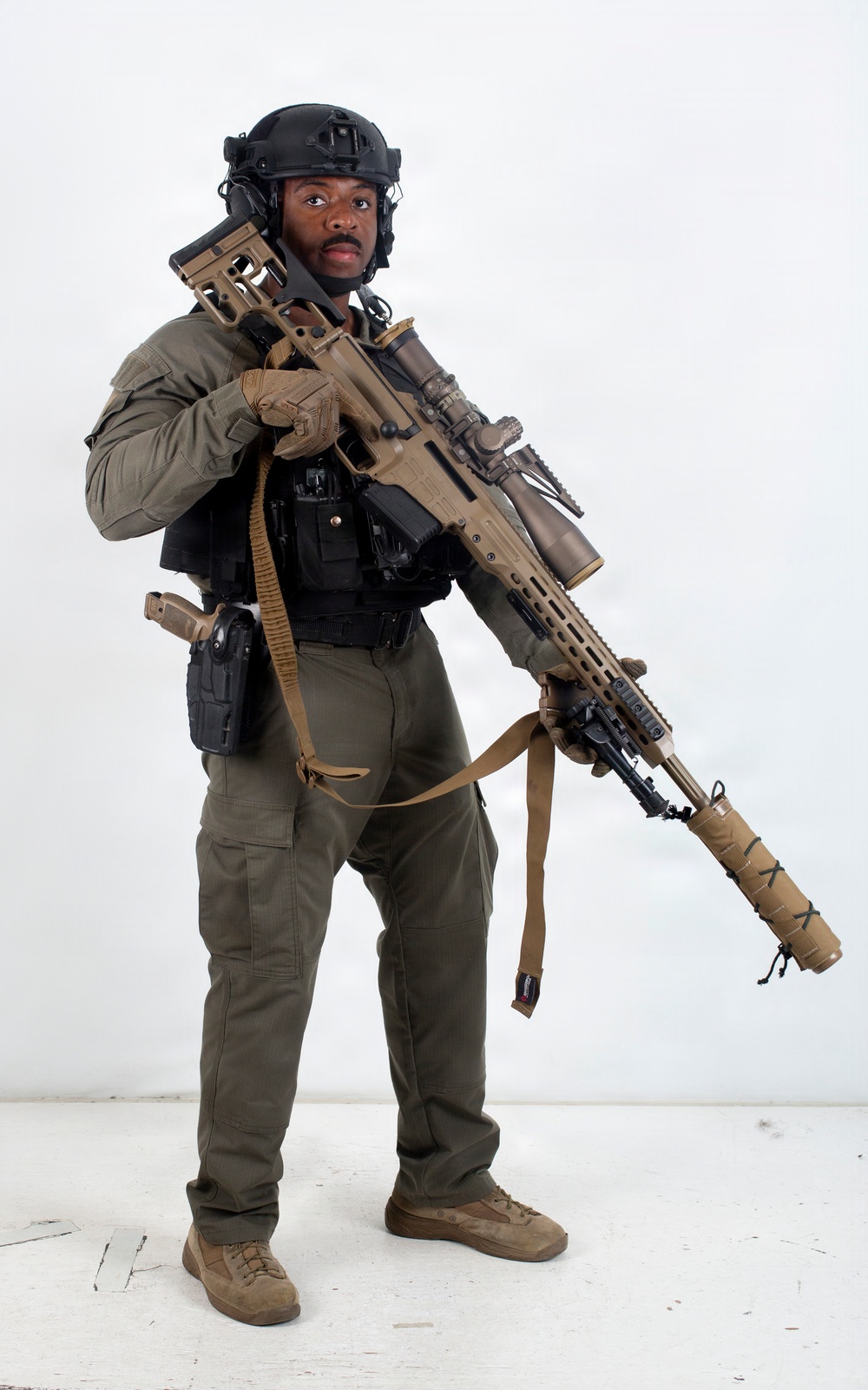 A person in full tactical/military-style gear is posed against a plain white background in what appears to be a studio setting. They are equipped with a large rifle featuring a suppressor, optic scope, and coyote-tan finish, along with a sidearm holster and tactical vest. The image appears to be a professional or promotional photograph.