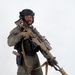 A person in full tactical/military-style gear is posed against a plain white background in what appears to be a studio setting. They are equipped with a large rifle featuring a suppressor, optic scope, and coyote-tan finish, along with a sidearm holster and tactical vest. The image appears to be a professional or promotional photograph.