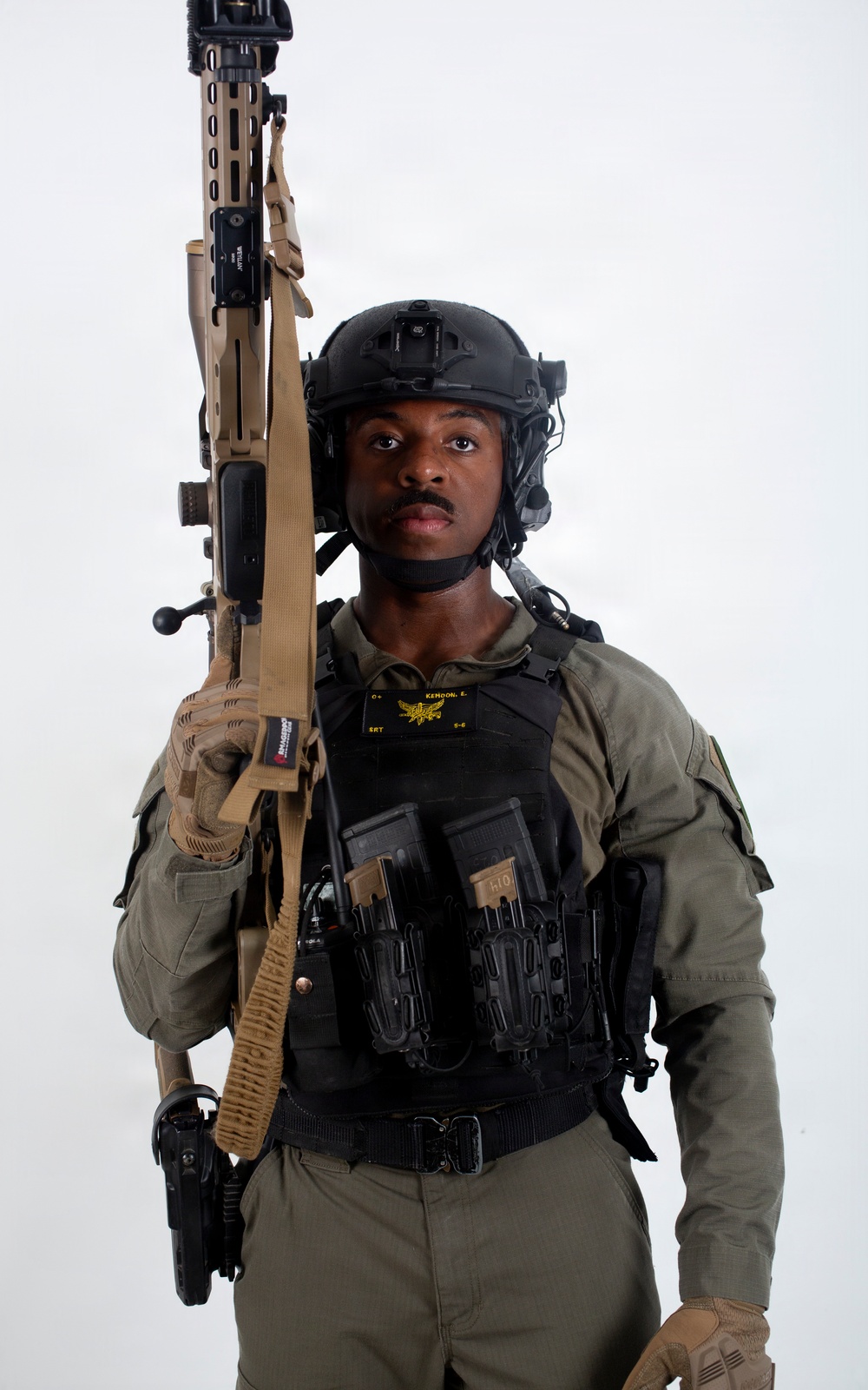 A tactical operator poses against a white studio background in full tactical gear. The individual is equipped with a plate carrier vest, ballistic helmet with communications equipment, and holds a tan/FDE-colored rifle with a sling. The image appears to be a professional portrait or official photo, likely law enforcement or military.