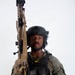 A tactical operator poses against a white studio background in full tactical gear. The individual is equipped with a plate carrier vest, ballistic helmet with communications equipment, and holds a tan/FDE-colored rifle with a sling. The image appears to be a professional portrait or official photo, likely law enforcement or military.