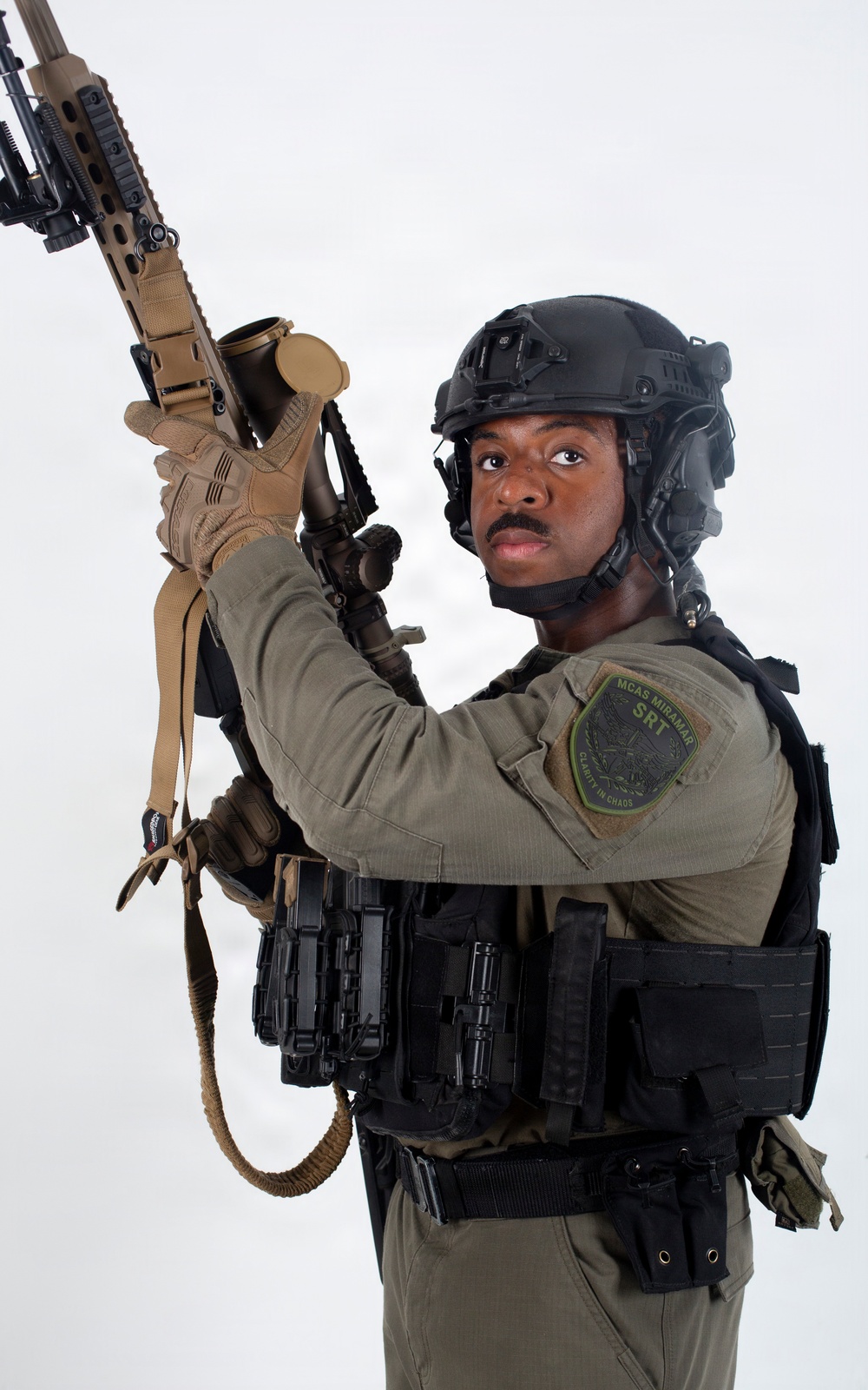 A uniformed security or military personnel member is posed against a white background holding a rifle upward. The individual is wearing full tactical gear including a ballistic helmet, plate carrier vest, and a patch identifying 'MCAS Miramar SRT' (Special Reaction Team). The image appears to be a professional portrait or promotional photograph.