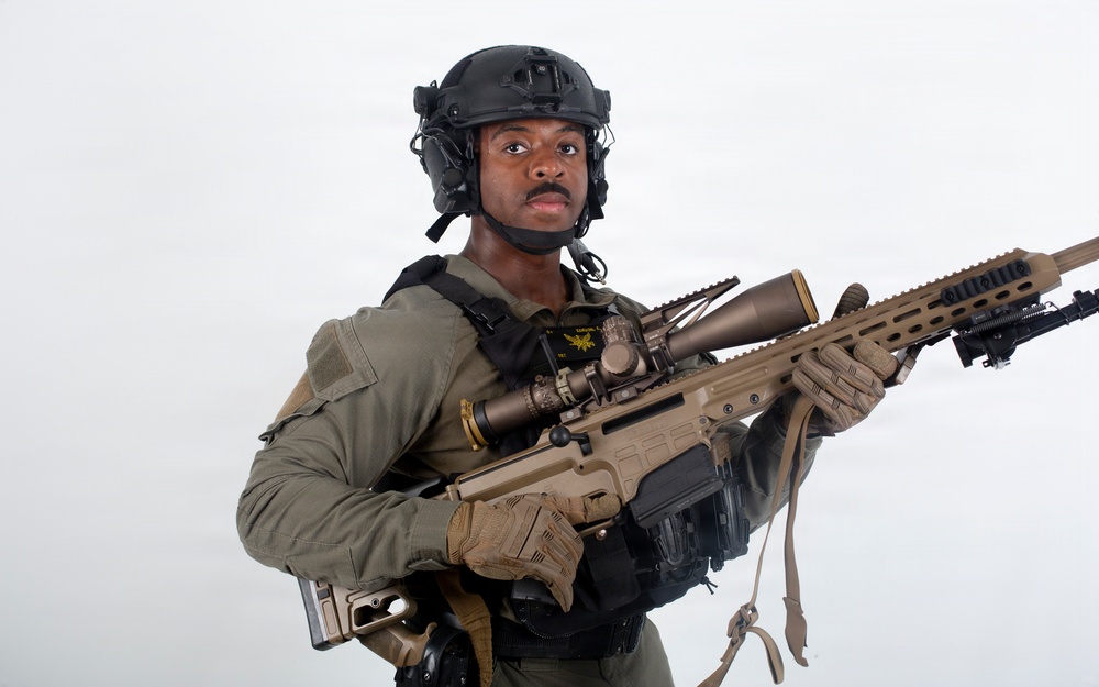 A military or law enforcement personnel is photographed against a plain white/grey background in what appears to be a professional or promotional setting. The individual is equipped with full tactical gear including a helmet with communications equipment and is holding a modern precision rifle. The image is well-lit and studio-quality.