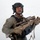 MCAS Marines pose for content 2026 A military or law enforcement personnel is photographed against a plain white/grey background in what appears to be a professional or promotional setting. The individual is equipped with full tactical gear including a helmet with communications equipment and is holding a modern precision rifle. The image is well-lit and studio-quality.