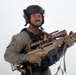 A military or law enforcement personnel is photographed against a plain white/grey background in what appears to be a professional or promotional setting. The individual is equipped with full tactical gear including a helmet with communications equipment and is holding a modern precision rifle. The image is well-lit and studio-quality.