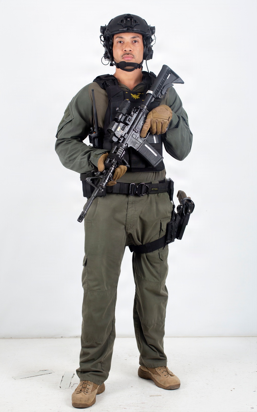 A male individual in full tactical gear is posed against a plain white background in what appears to be a professional studio photograph. He is equipped with military/law enforcement-style equipment including body armor, helmet, and weapons. The image appears to be a reference or documentation photograph.