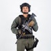 A male individual in full tactical gear is posed against a plain white background in what appears to be a professional studio photograph. He is equipped with military/law enforcement-style equipment including body armor, helmet, and weapons. The image appears to be a reference or documentation photograph.