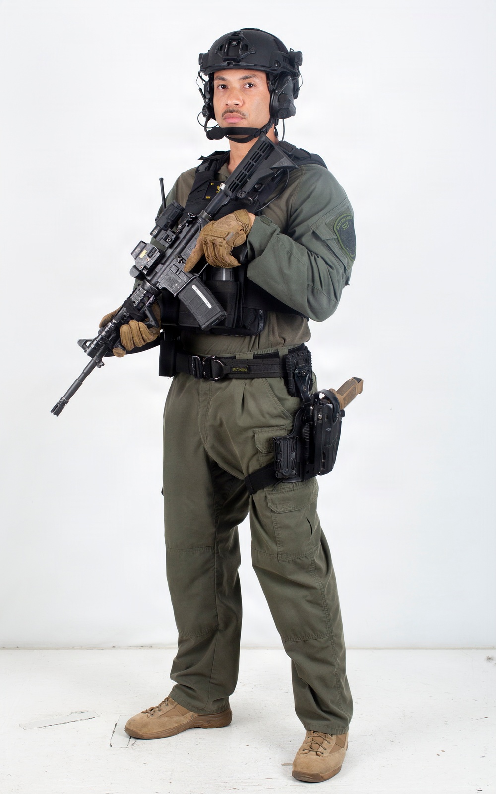 A uniformed tactical law enforcement officer stands in a studio setting against a white background. He is fully equipped with tactical gear, body armor, communications equipment, and is holding an assault-style rifle. A patch on his left sleeve and a badge on his chest suggest law enforcement affiliation, likely a SWAT or SRT (Special Response Team) unit.