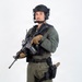 A uniformed tactical law enforcement officer stands in a studio setting against a white background. He is fully equipped with tactical gear, body armor, communications equipment, and is holding an assault-style rifle. A patch on his left sleeve and a badge on his chest suggest law enforcement affiliation, likely a SWAT or SRT (Special Response Team) unit.