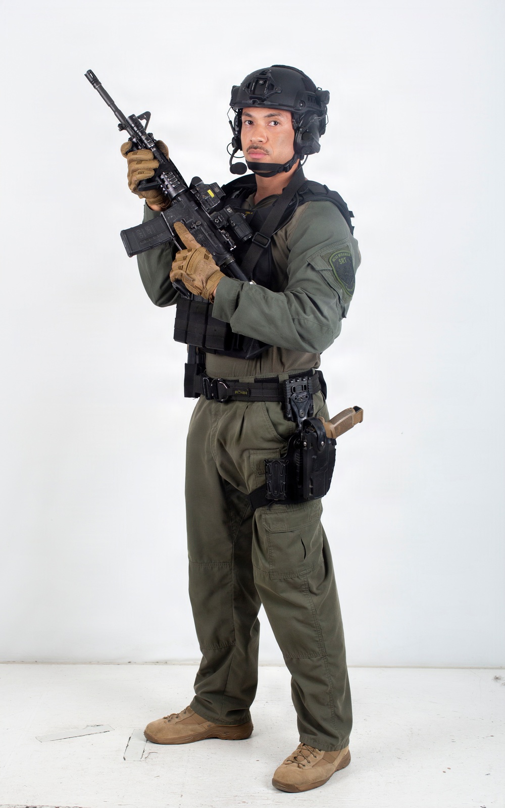 A male individual in full tactical law enforcement or military-style gear is posed against a plain white studio background. He is holding an assault-style rifle in a ready position and is equipped with extensive tactical equipment. The image appears to be a professional studio photograph, likely for promotional or documentation purposes.